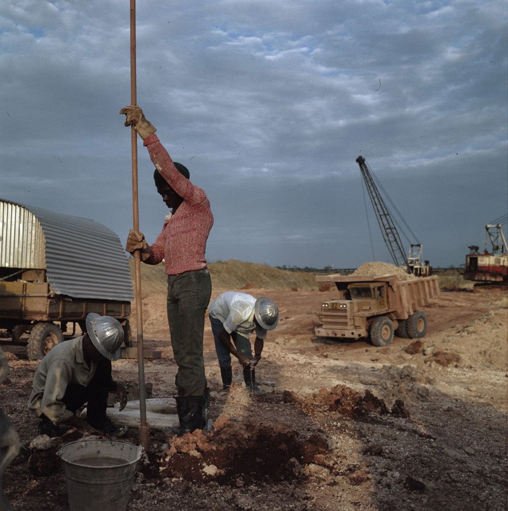 A worker stands using a long pole as others wearing hard hats pend down and sift through dirt. A dump truck and heavy equipment are seen behind them; heavy gray clouds loom overhead.