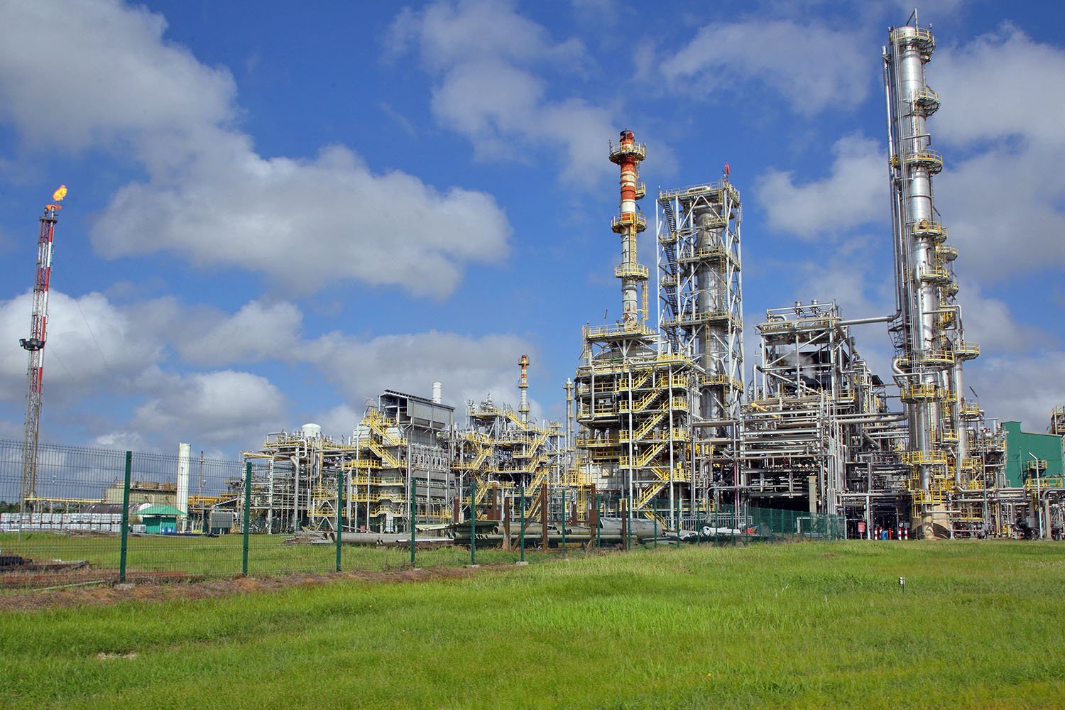 The towers of an oil refinery stretch up against a blue sky. The grass in the foreground is lush and green.