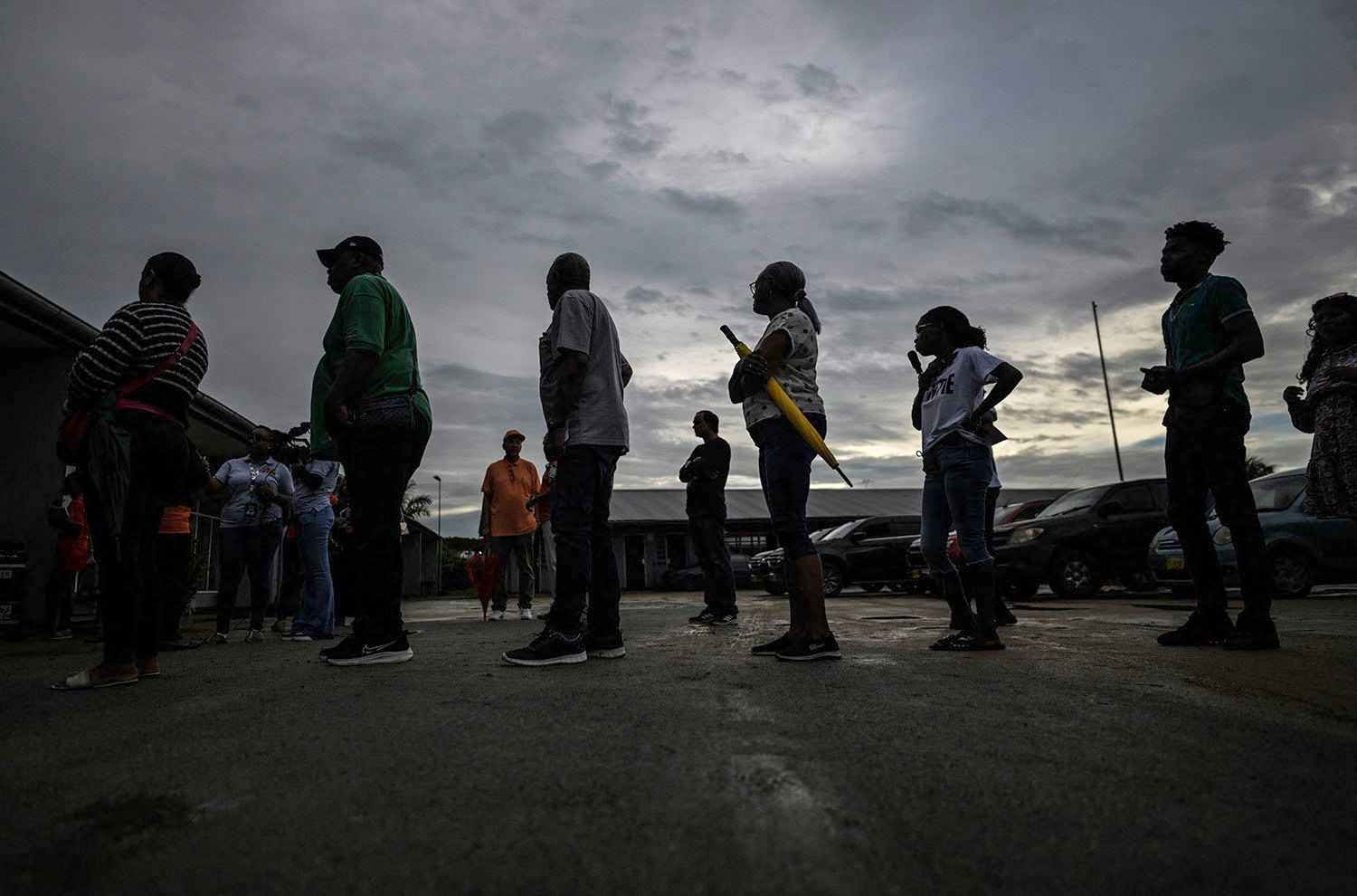 People are seen from the side as they stand on a stretch of pavement in a line against a dim gray sky.