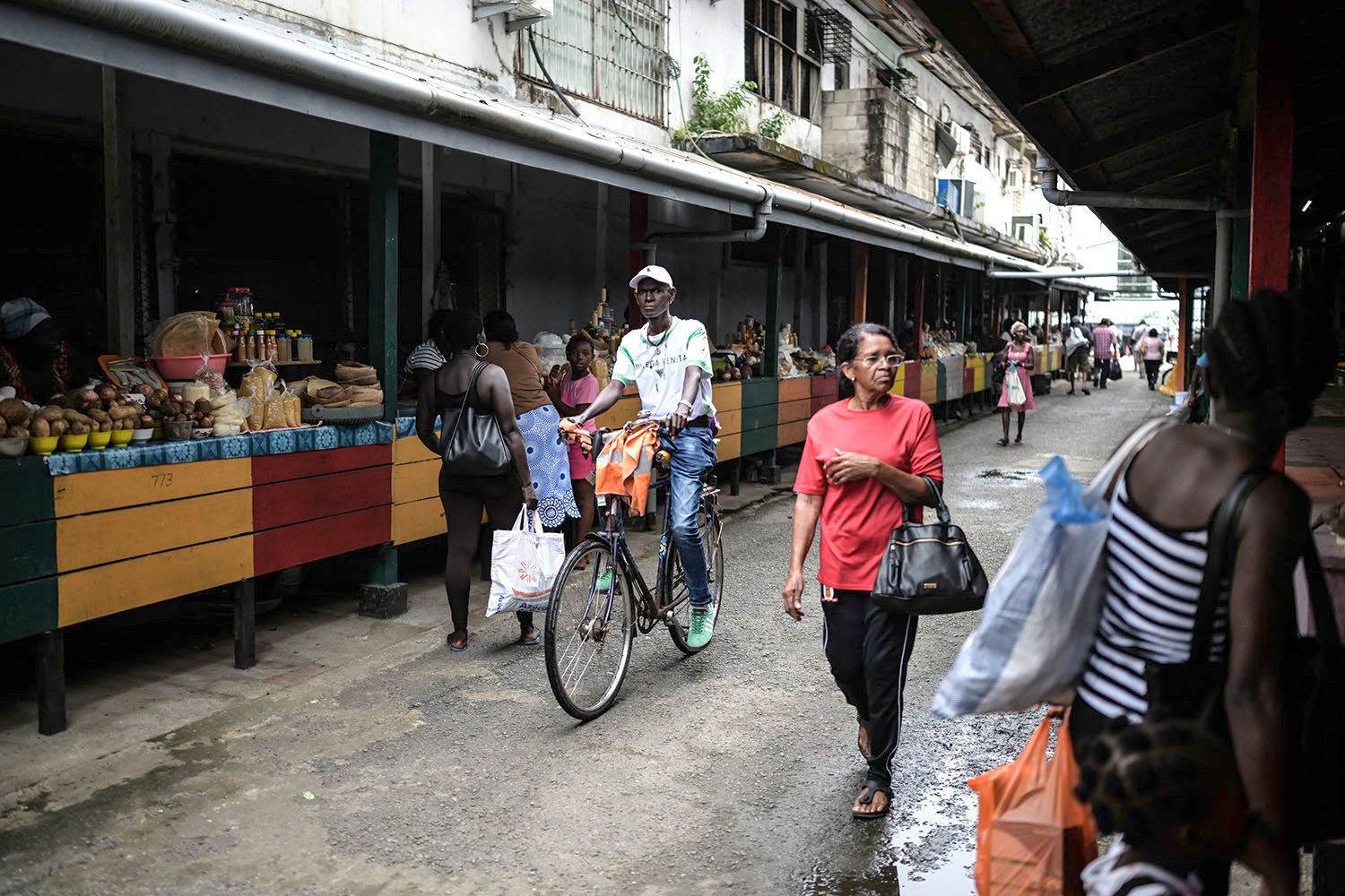 A man rides a bike down a street in front of a marketplace covered with an awning. Other passersby include several women with a shopping bags.