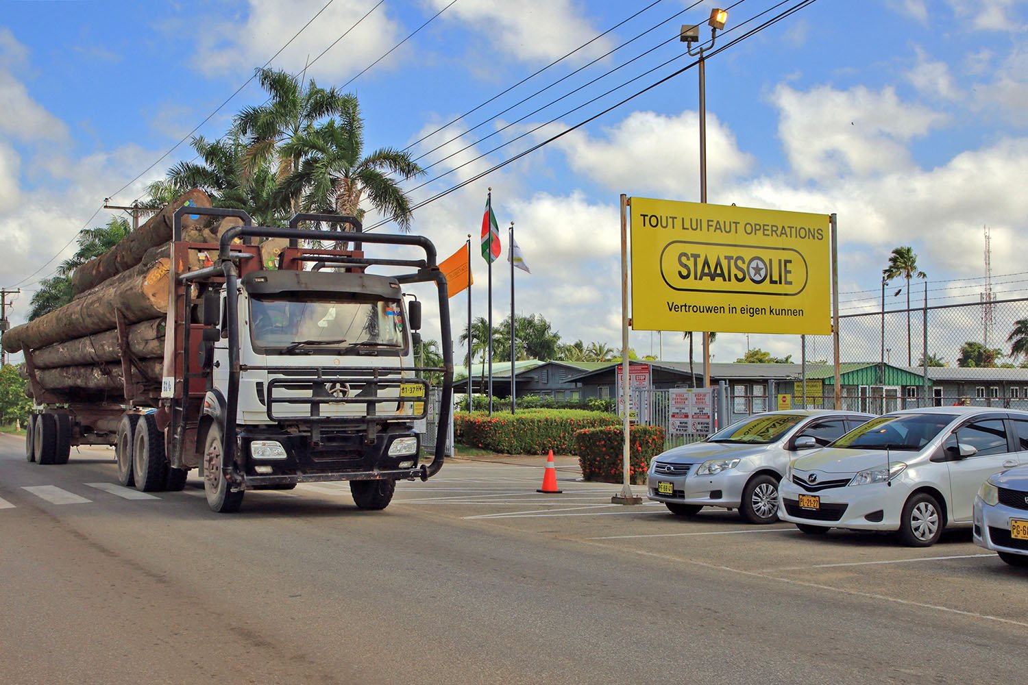 A truck carrying palm trees drives past a sign for Staatsolie. Power lines are above against a blue sky.