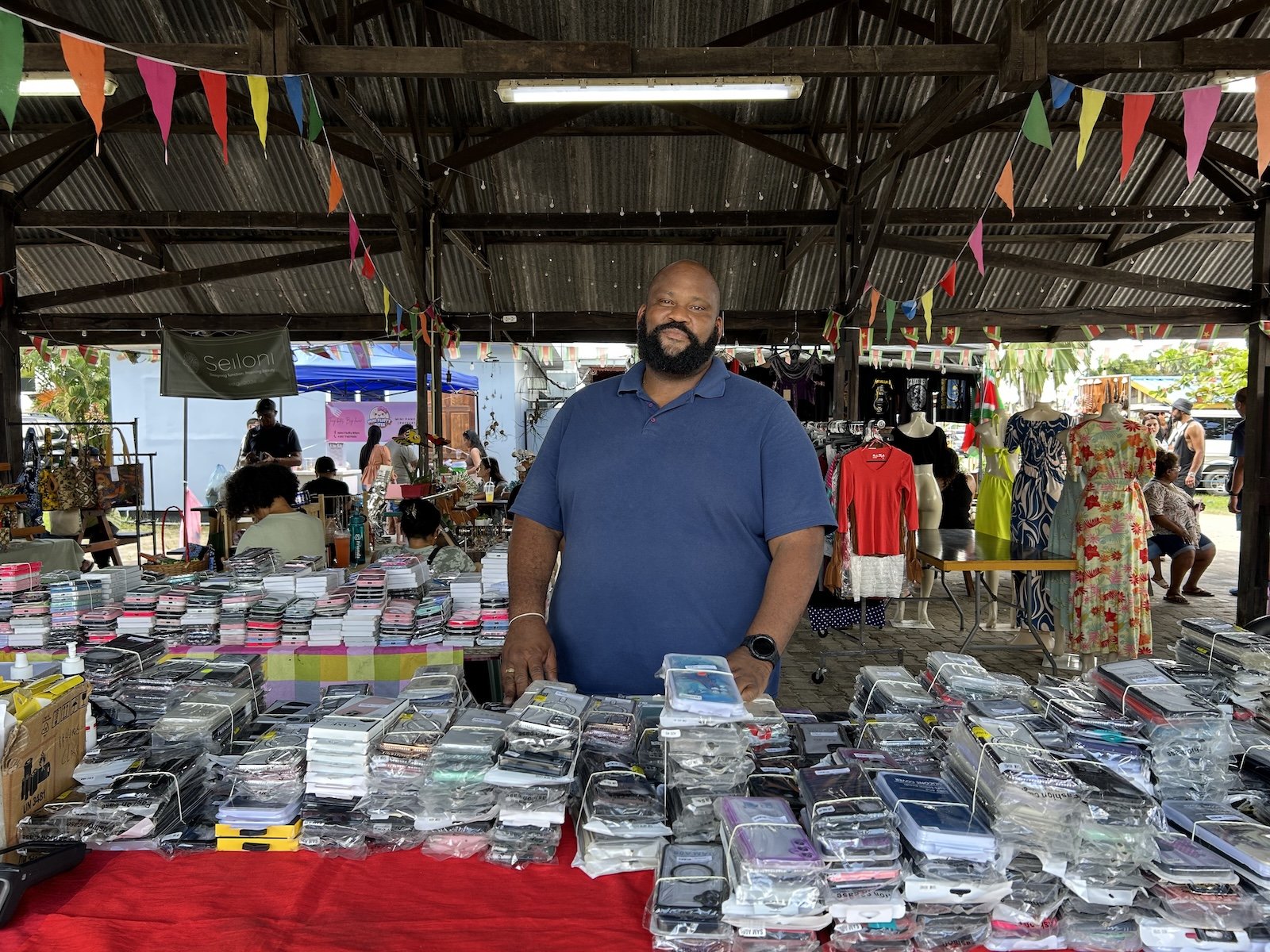 A man in a blue polo shirt stands in a book behind a table full of stacks of phone accessories and other products.