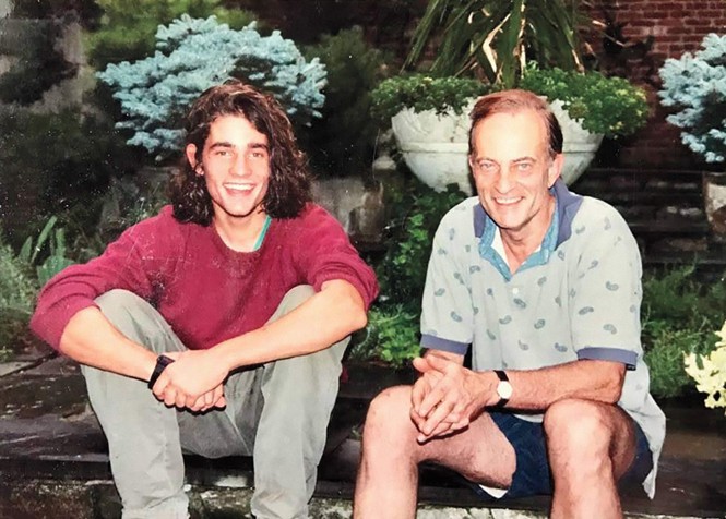 candid photo of smiling young man with shoulder-length brown hair and arms clasped around knees sitting next to older man wearing polo and shorts at edge of garden