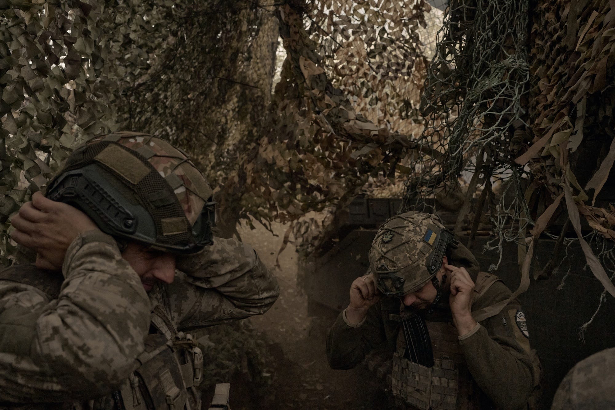 Two men in camouflage gear cover their ears under netting