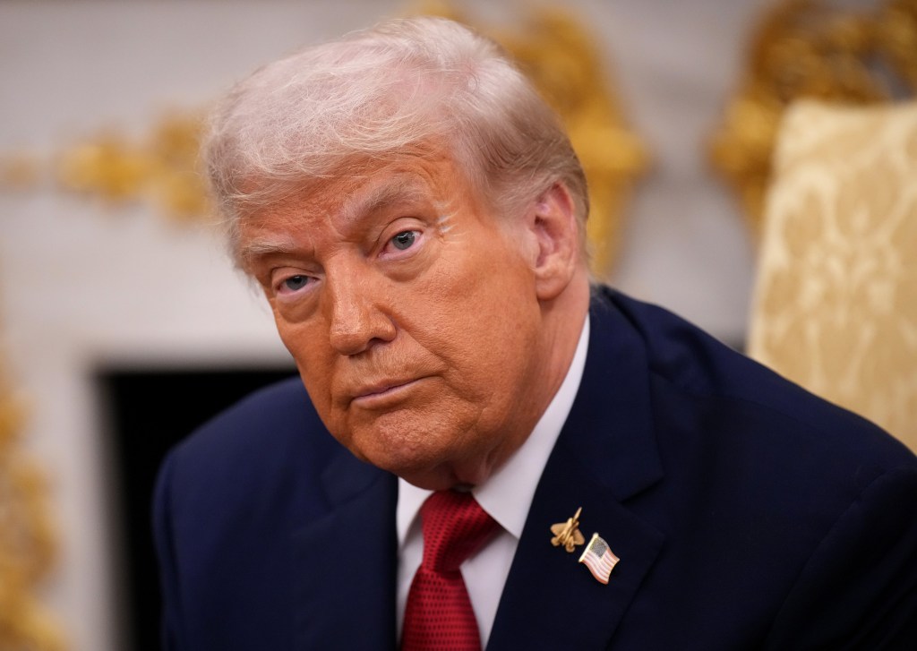 Donald Trump looking at the camera, wearing a blue suit, red tie, and a US flag pin with an F-35 jet pin.