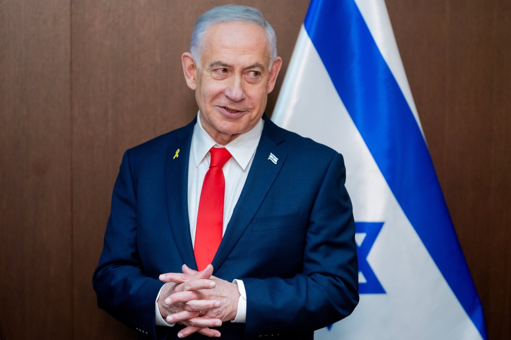 Israel's Prime Minister Benjamin Netanyahu smiles, wearing a suit with a yellow ribbon and an Israeli flag pin, with an Israeli flag in the background.
