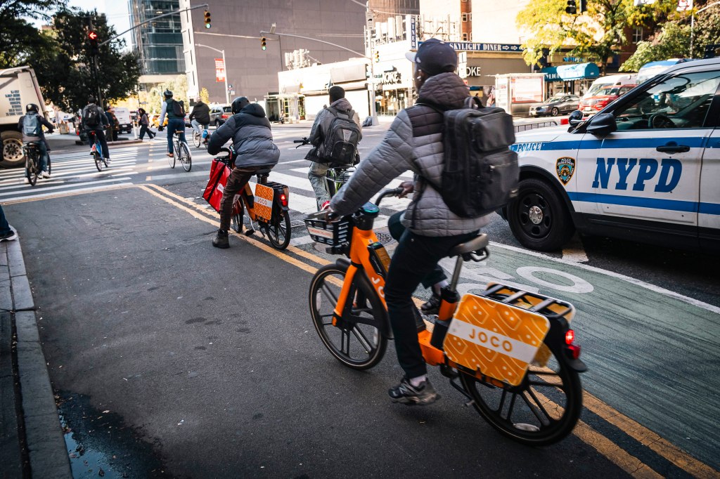 Cyclists on Joco e-bikes ride past an NYPD patrol car in Manhattan.