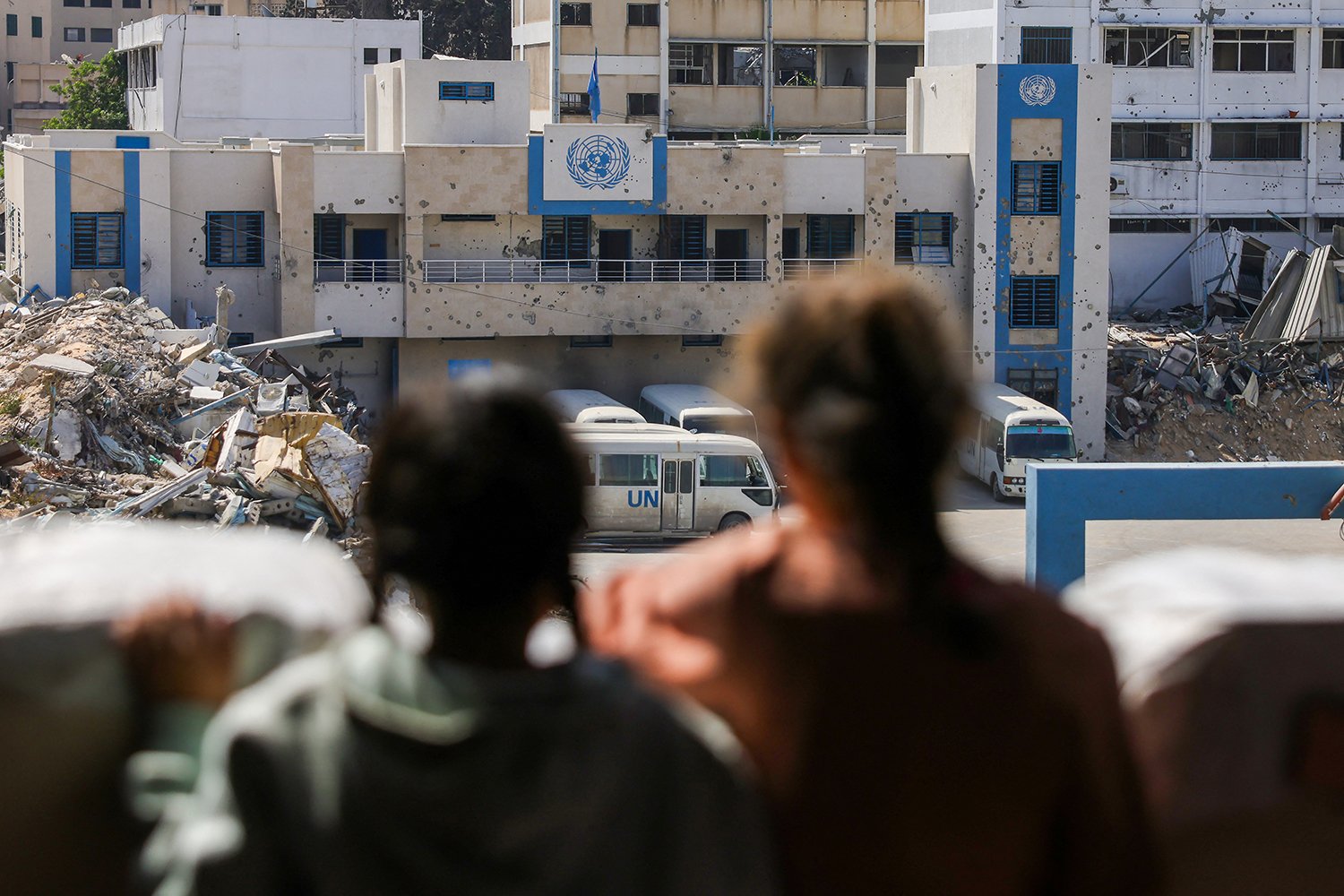 Two children are seen out of focus in the foreground looking at the pock-marked, heavily damaged building with the U.N. logo on it.