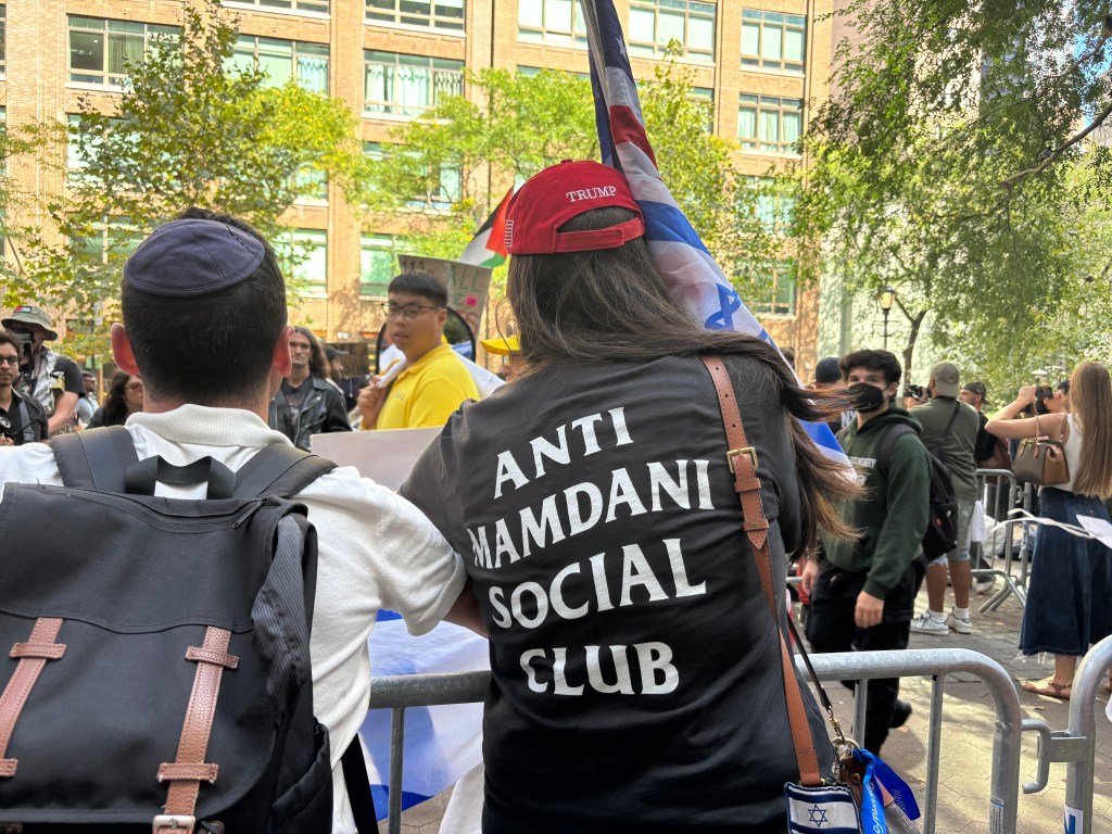 Back of a person wearing a red MAGA hat and a black t-shirt that says “ANTI MAMDANI SOCIAL CLUB” at a protest.