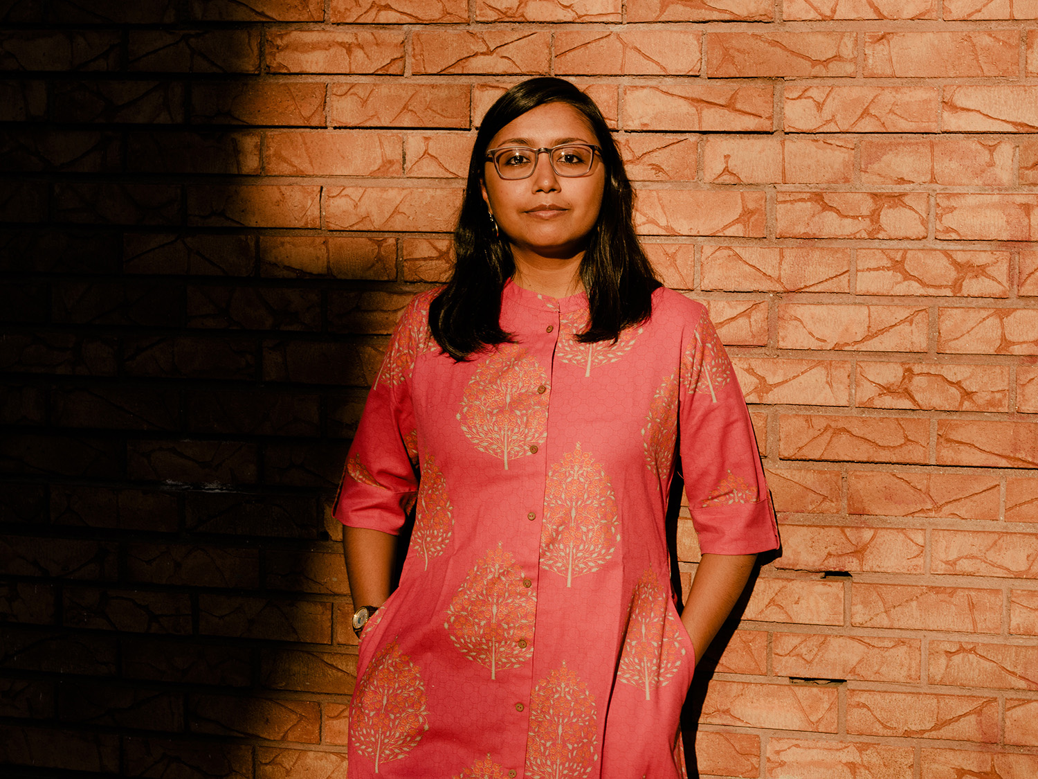 A woman wearing glasses and a patterned tunic stands against a brick wall.
