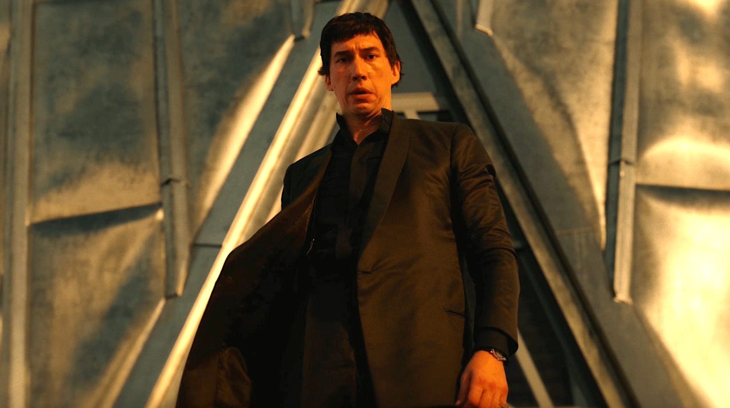 A man with dark hair in a black suit looks down at the camera with a metal wall behind him.