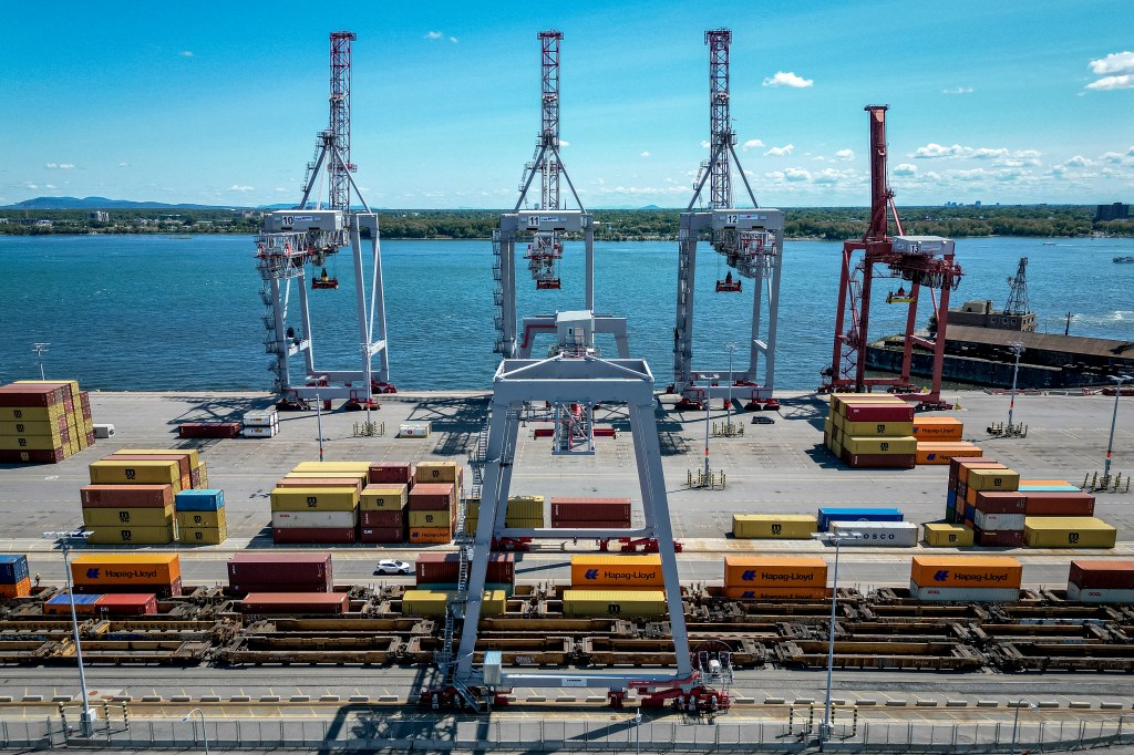 Shipping containers stacked at the Port of Montreal in Quebec, Canada, with large gantry cranes overlooking them and the water.