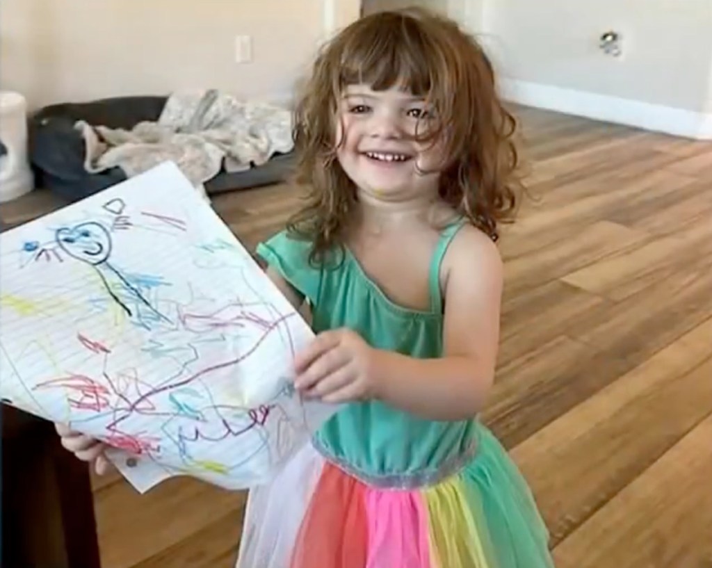 Parker Scholtes, a young girl with brown curly hair, smiling and holding a drawing.
