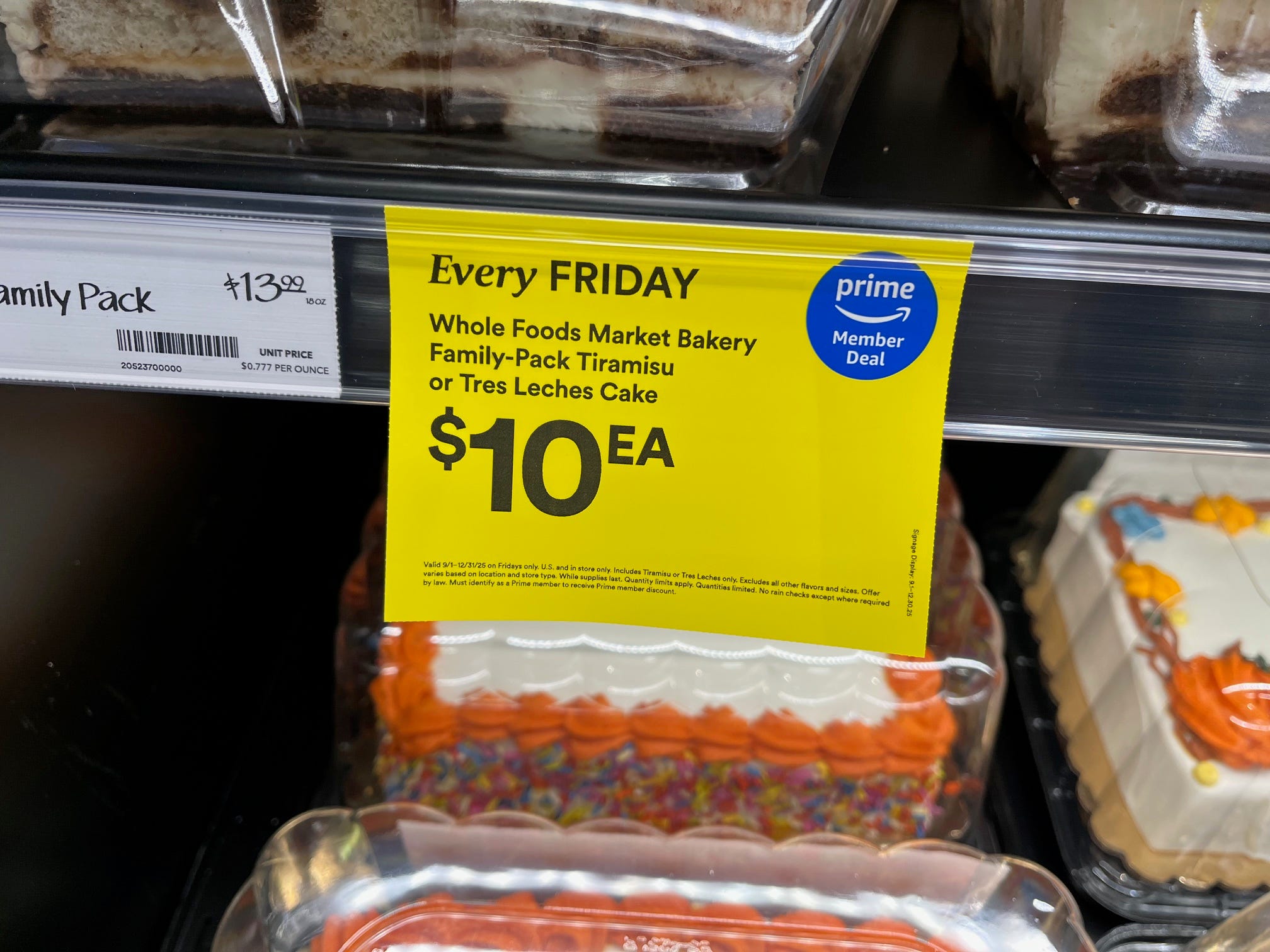 A yellow shelf tag at Whole Foods Daily Shop shows a discounted price of $10 on Fridays for a family pack of Tiramisu or Tres Leches cake.