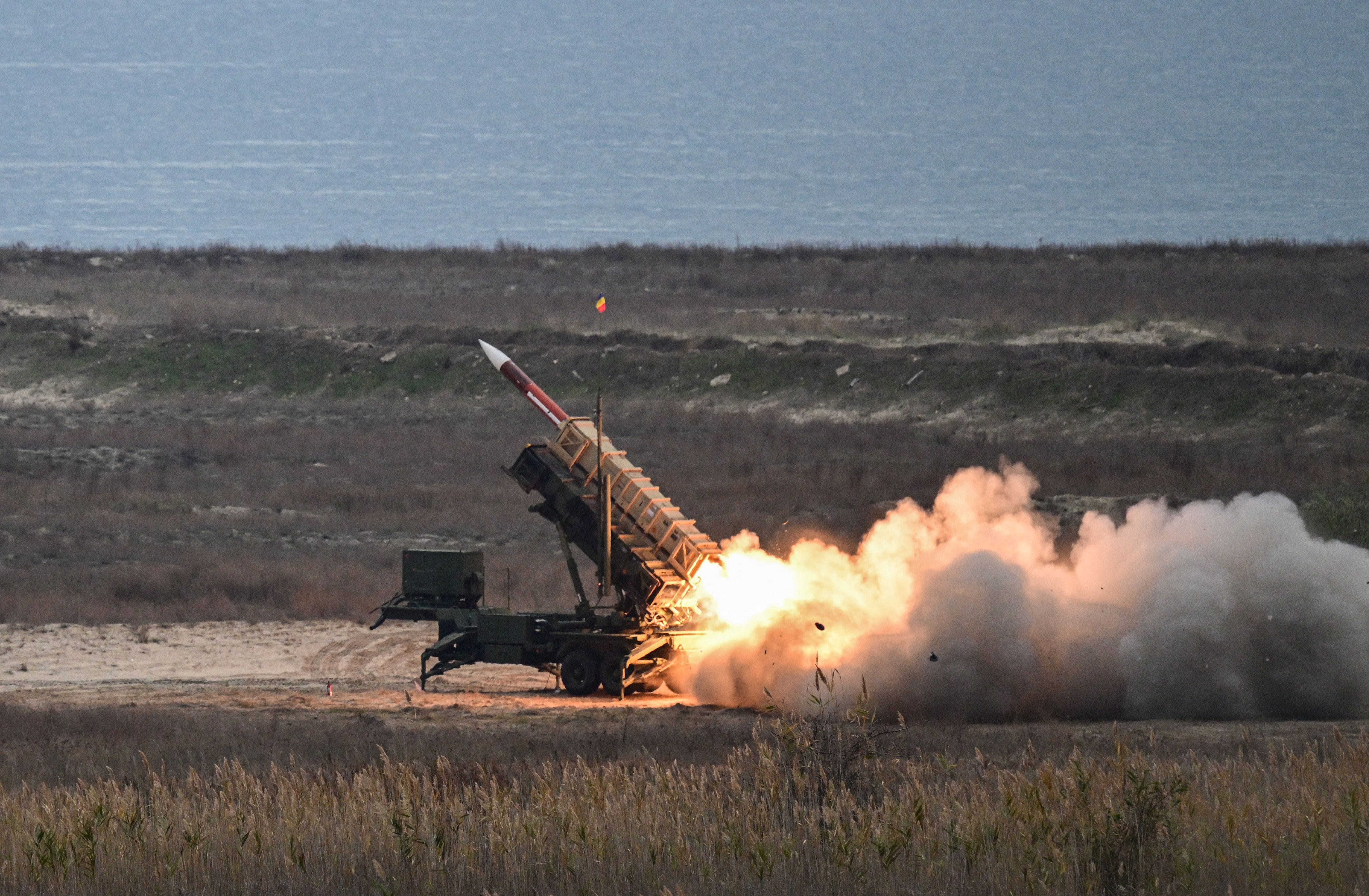 A missile is launched from a large system with a body of water in the background