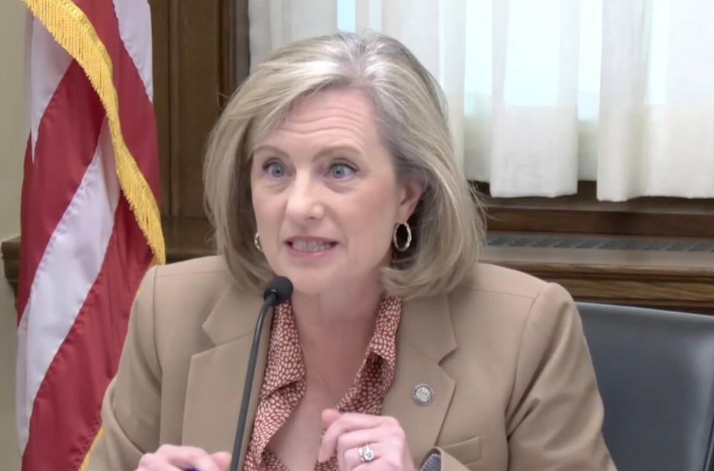 A woman in a tan blazer and patterned shirt speaks into a microphone.