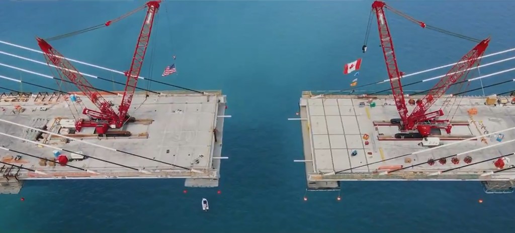 Aerial view of a bridge under construction, with American and Canadian flags flying from cranes.