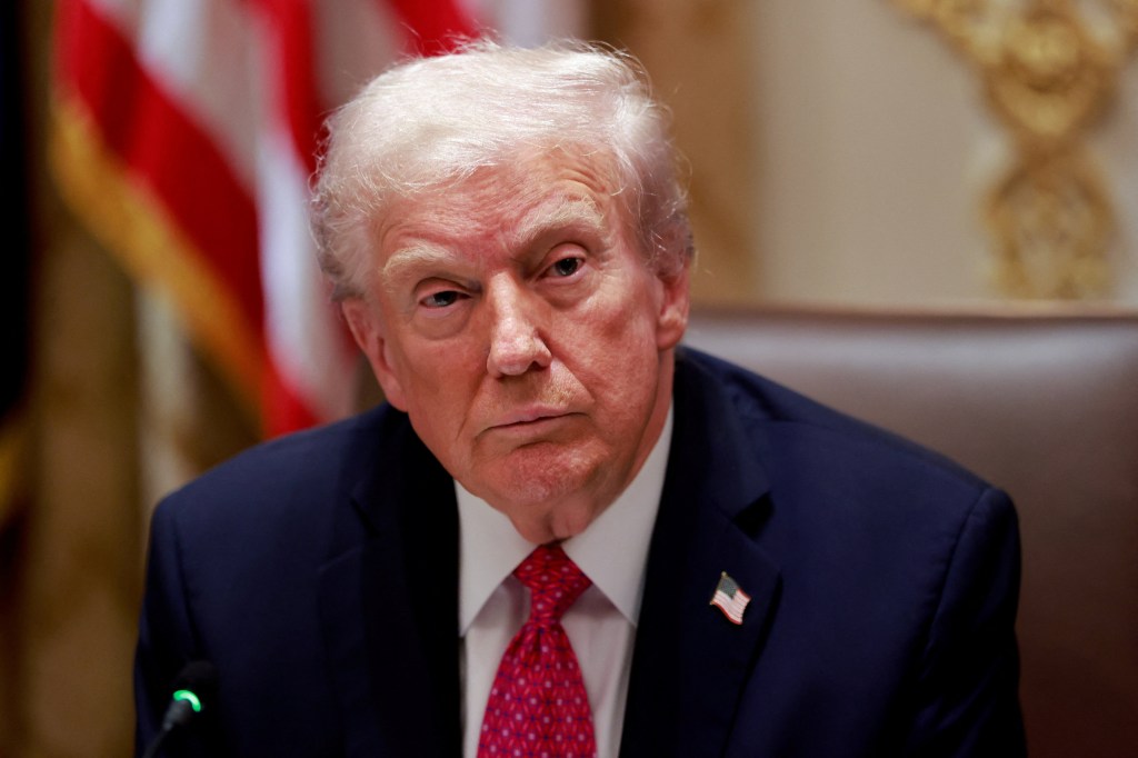 President Donald Trump in a dark suit and red patterned tie, with an American flag lapel pin, looks directly at the camera.