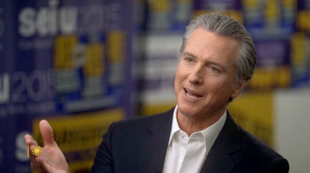 California Governor Gavin Newsom, wearing a suit, speaking during an interview with a blurred SEIU 2020 background.
