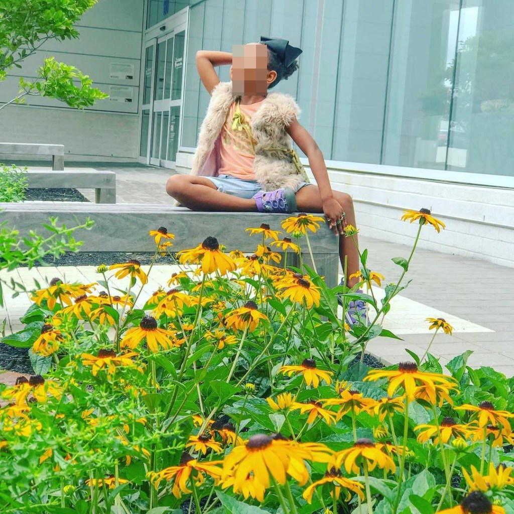 Divine, daughter of Karizma Dabney, sitting on a bench behind a bed of yellow flowers.