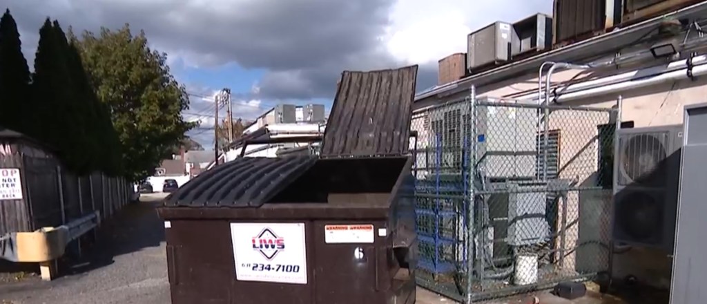 A dumpster behind a closed grocery store, with its lid partially open, next to a chain-link fence enclosing industrial equipment.