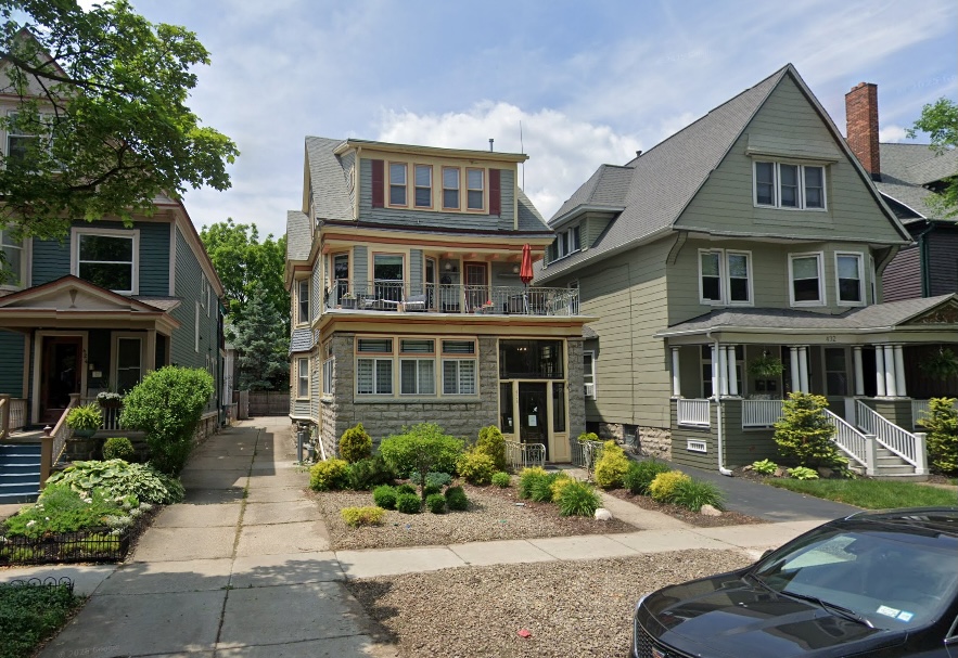 The property at 428 Norwood Ave, Buffalo, NY, is a three-story house with a stone base and light blue siding, featuring a second-story balcony with a red umbrella, flanked by similar residential buildings and a black car parked in the foreground.