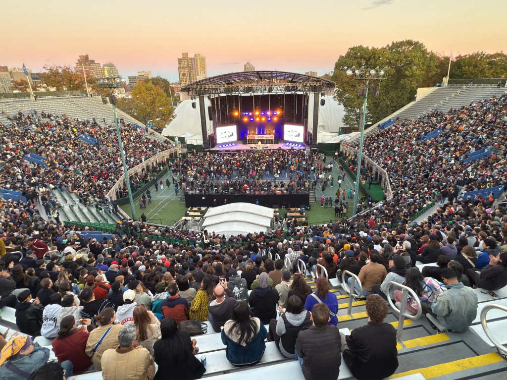 General view of a rally in Queens with a large crowd gathered in stadium seating facing a brightly lit stage.