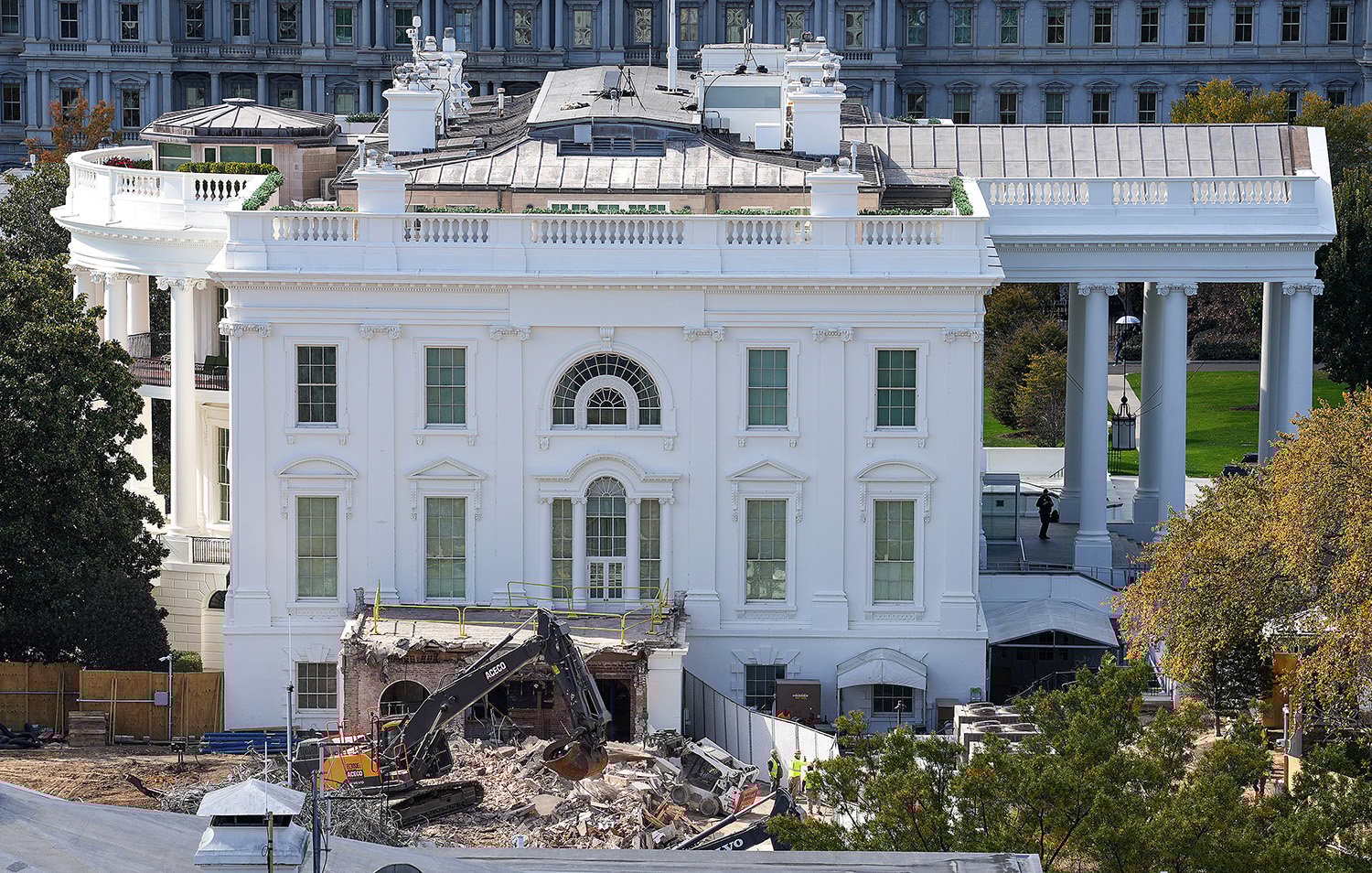 An excavator sits atop a pile of rubble on a construction site on the side of the White House. The grass around the building is vivid green, and the leaves of the trees around it are beginning to run yellow.