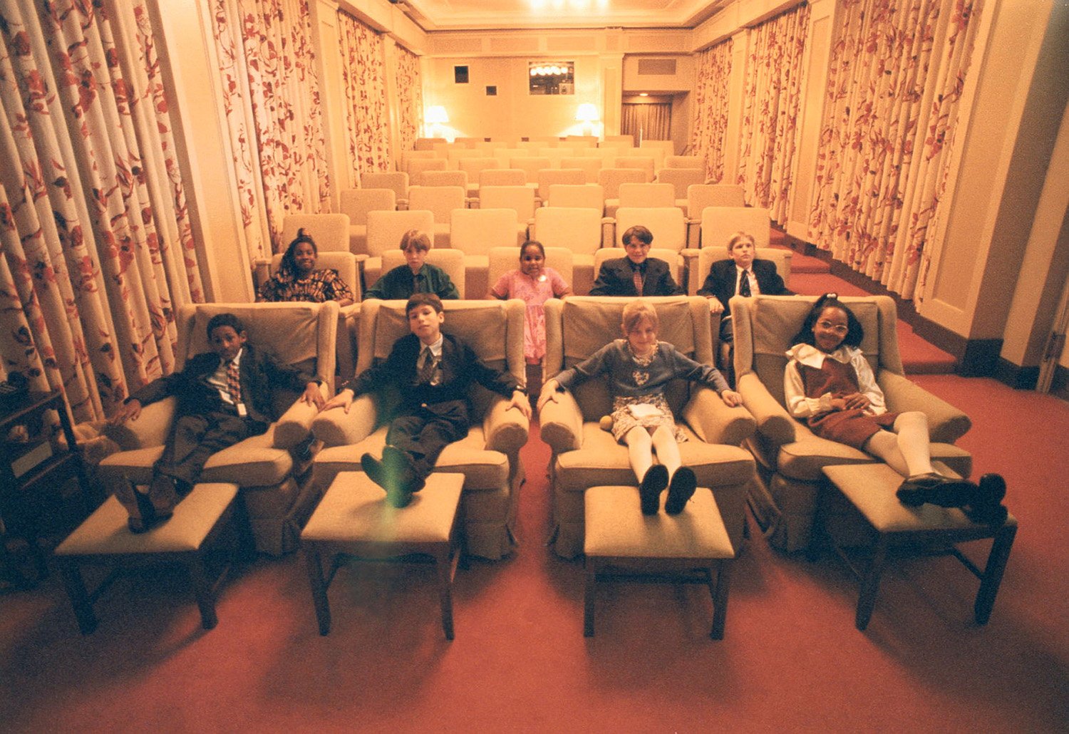 Nine children are seated in the front two rows of a curtain-draped movie room with multiple rows of plush chairs. The children are dressed in suits and dresses with their feet resting up on stools.
