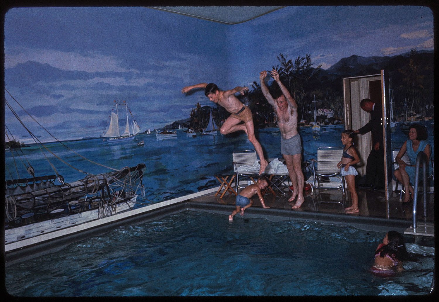 A shirtless man and boy jump into a pool. Children swim or stand on the deck nearby, with a woman in a blue dress seated behind them and a man in a suit standing in a doorway. The walls of the pool area are painted with a nautical scene.