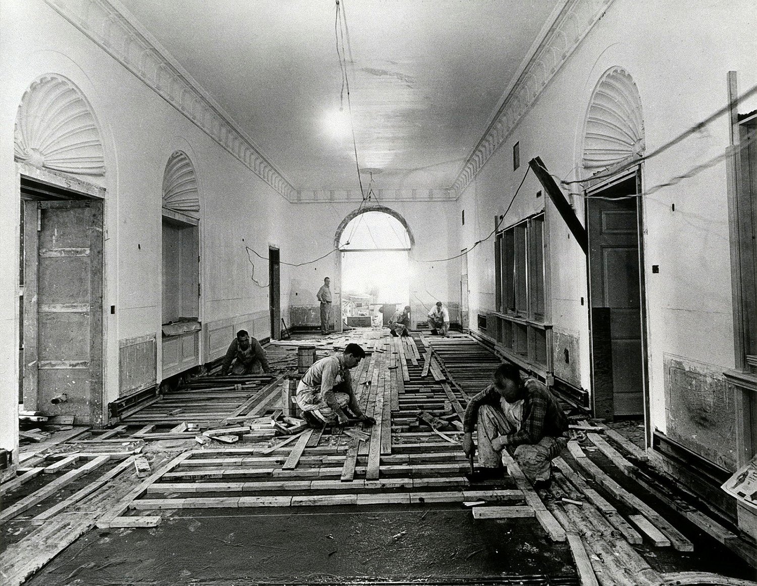 Workers kneel as they lay flooring tiles in a disheveled hallway in the White House with high ceilings and tall arched doorways. A bare lightbulb hangs from an exposed wiring system overhead.