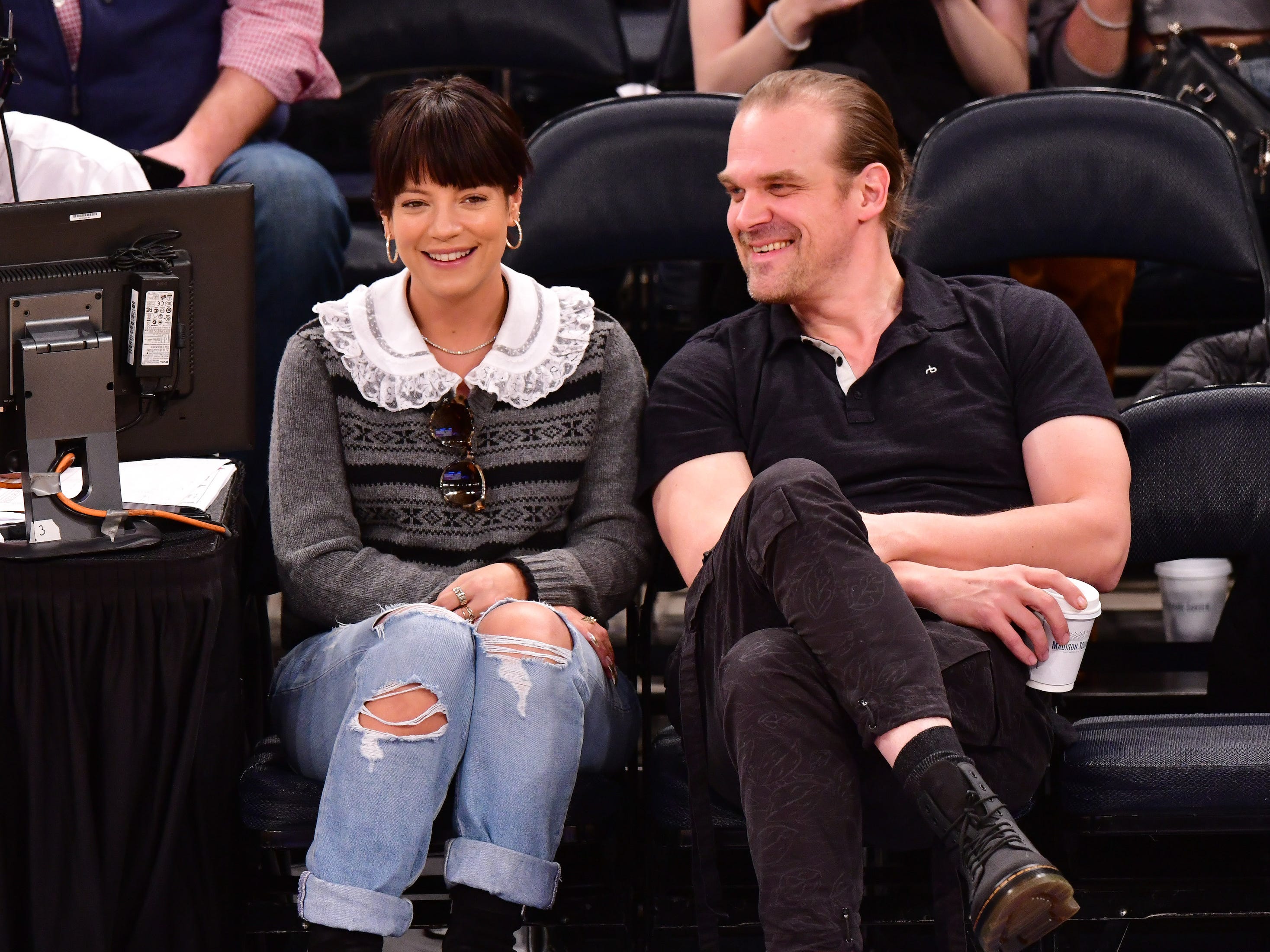 Lily Allen, in a gray sweater, white blouse, and jeans, sits court side at a New York Knicks game with David Harbour, wearing all black, in 2019.