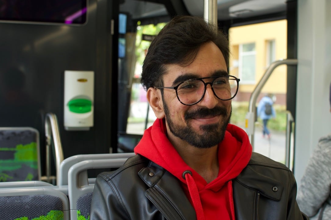 A bearded man in glasses with a black jacket over a red hoodie sits in a bus seat.