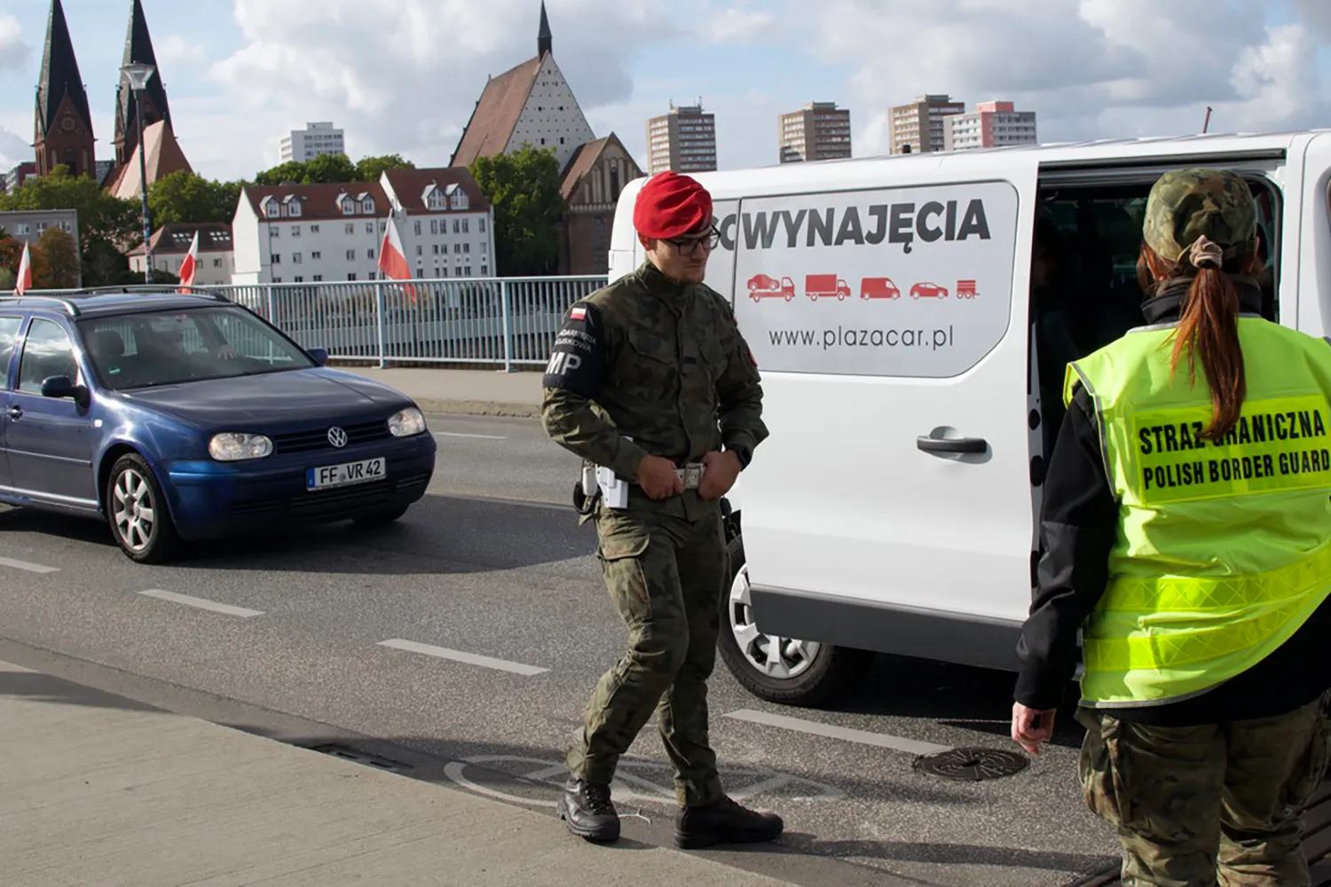 An officer in a red beret stands next to an open white van with Polish lettering on it. A person in a yellow reflective vest stands at right. Cars drove along the street and buildings and Polish flags are seen in the distance.