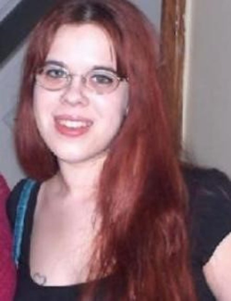 Deirdre Harm, with red hair and glasses, smiles slightly.