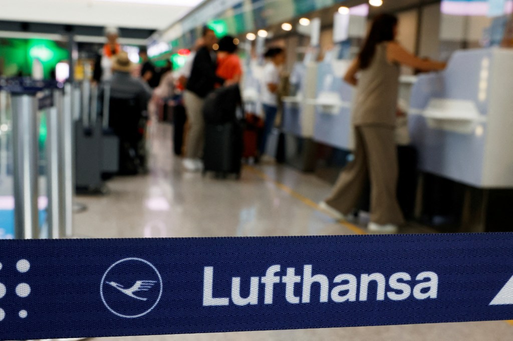 A blue barrier tape with the Lufthansa logo and name, separating the foreground from a blurry background of people at an airport check-in.