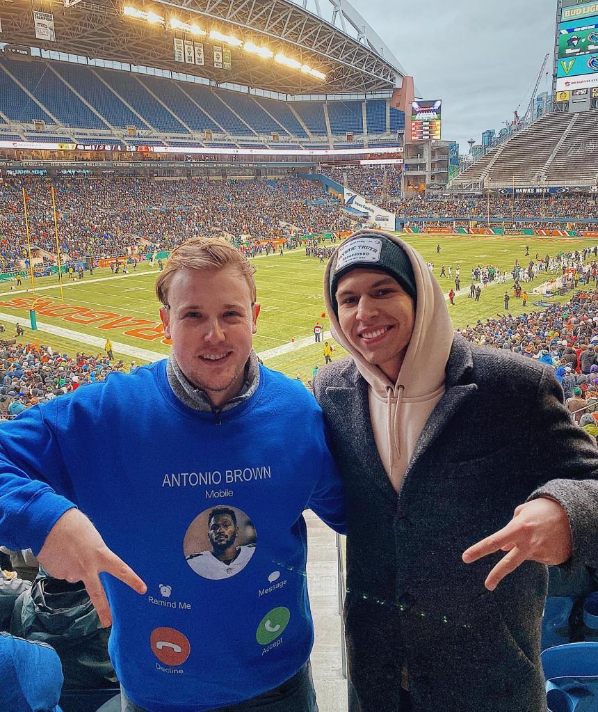 Matthew Cohn in a blue sweatshirt pointing at his shirt, posing with another man, smiling in a crowded football stadium.