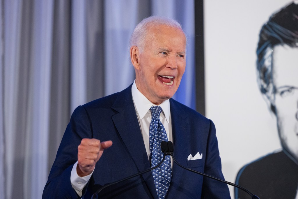 Former President Joe Biden speaking at a podium with a microphone, gesturing with his right hand, and smiling widely.
