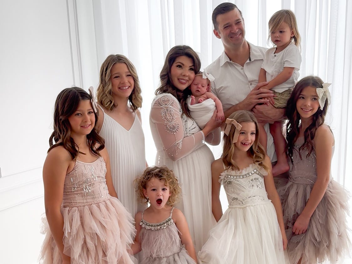 A family of nine poses for a photograph wearing white and pastel outfits.