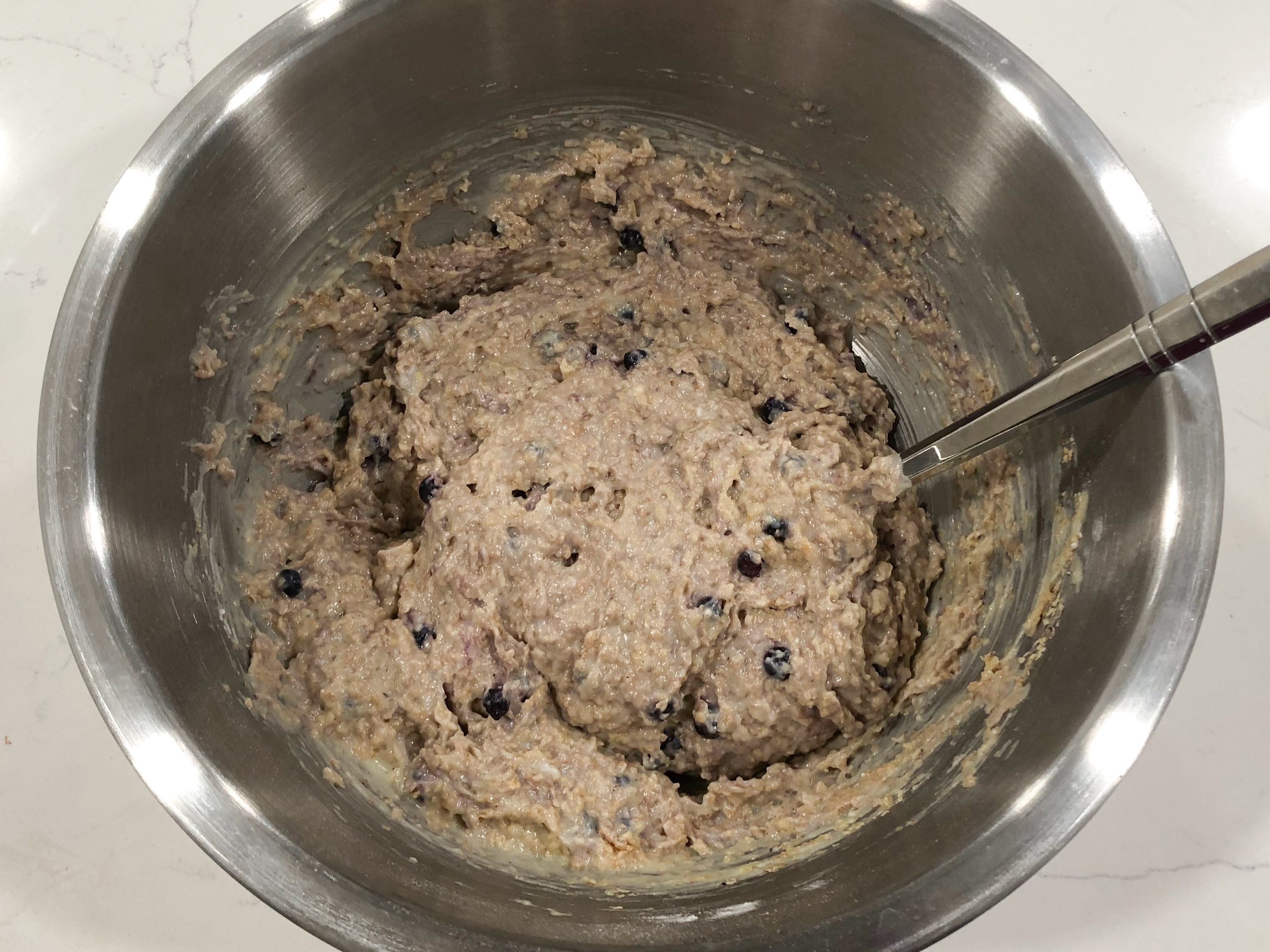 Muffin batter, which now includes blueberries and is a darker color, in a stainless-steel bowl with a spoon.
