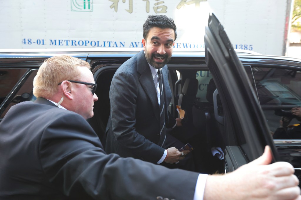 NYC 2025 Mayoral candidate Zohran Mamdani exits a vehicle, smiling at someone off-camera, while a man in a suit stands in the foreground.