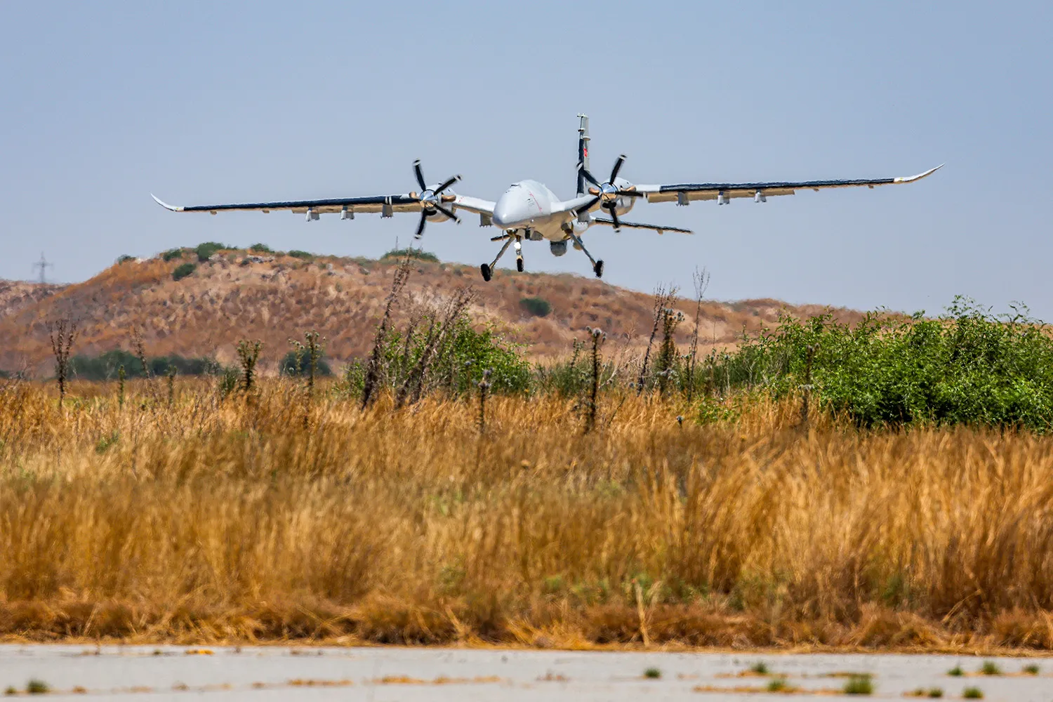 A drone is seen a few feet above the ground as it comes into land above a hillside covered with tall yellowed grass. The ground rises up to form sparsely covered hills on the horizon.