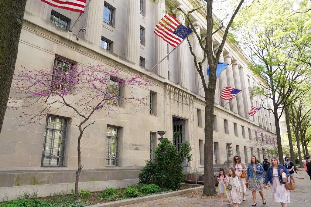 The Department of Justice building in Washington, D.C., with American flags flying and people walking along the sidewalk.