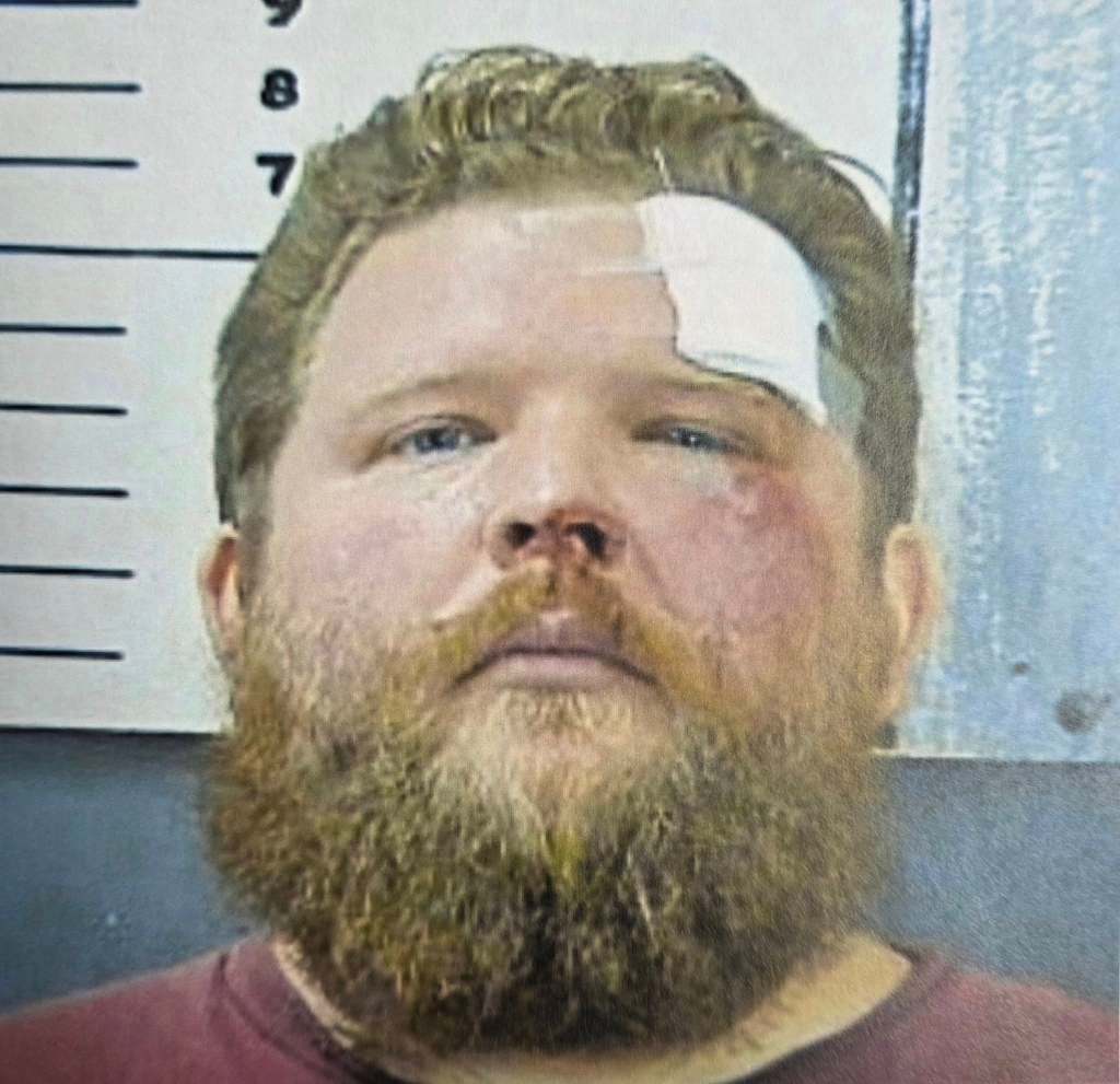 A mugshot of a man with a beard and a bandage on his forehead, bruises on his face.