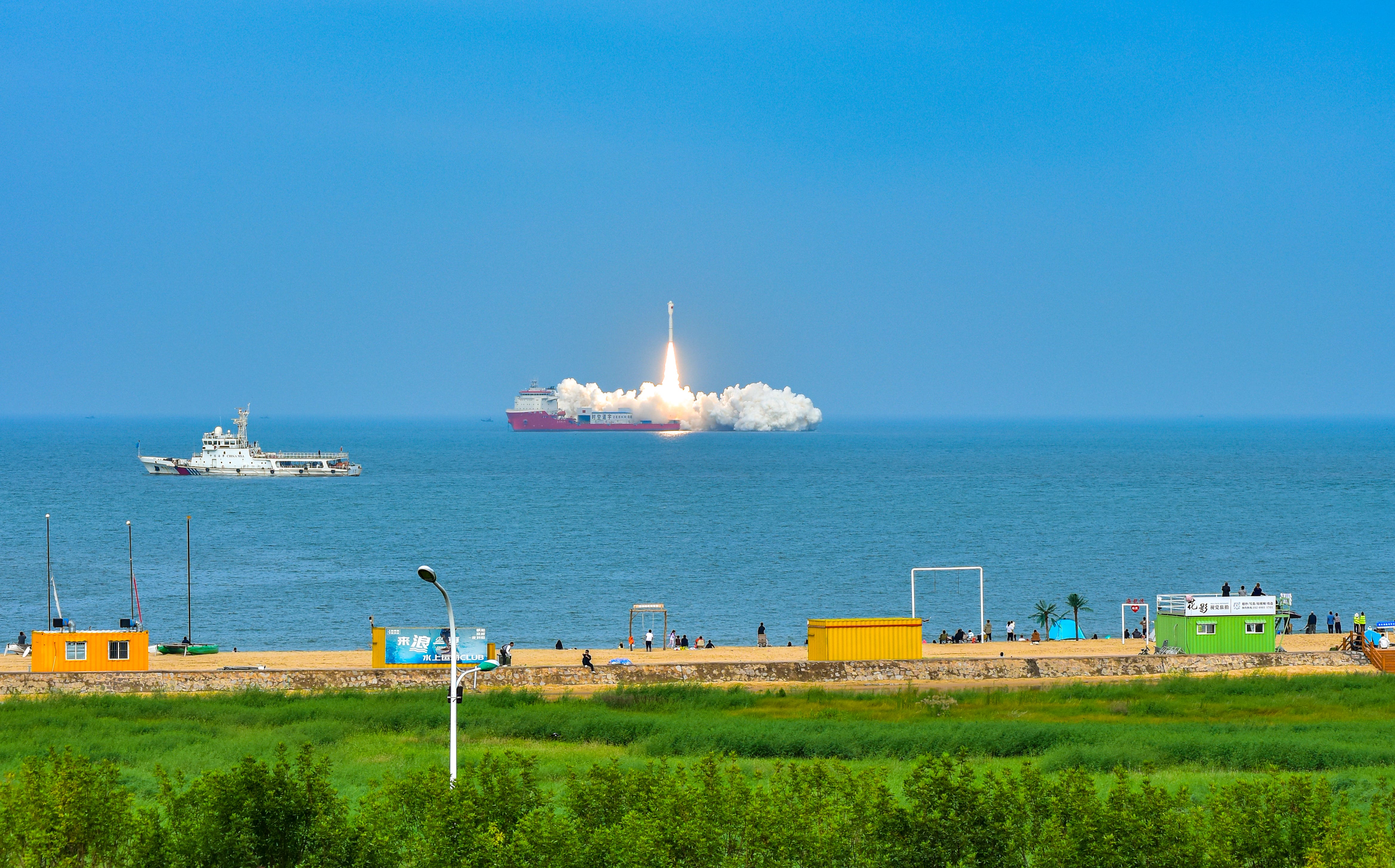 A Chinese rocket launches from a boat in the ocean, with the land and beach seen in the foreground of the picture and a clear blue sky in the background.