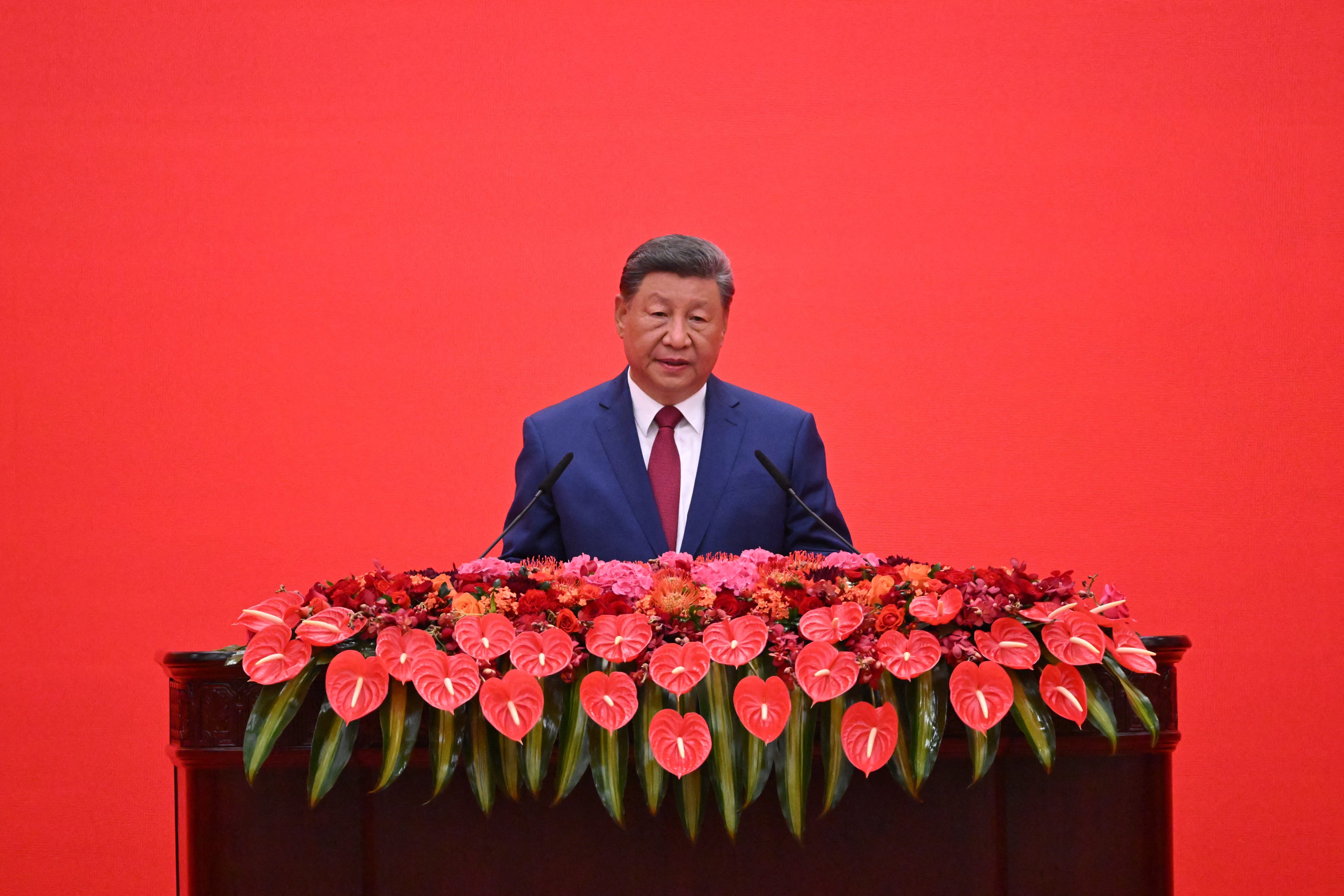 Chinese Communist Party leader Xi Jinping stands at a podium with a red background and red flowers on the podium.