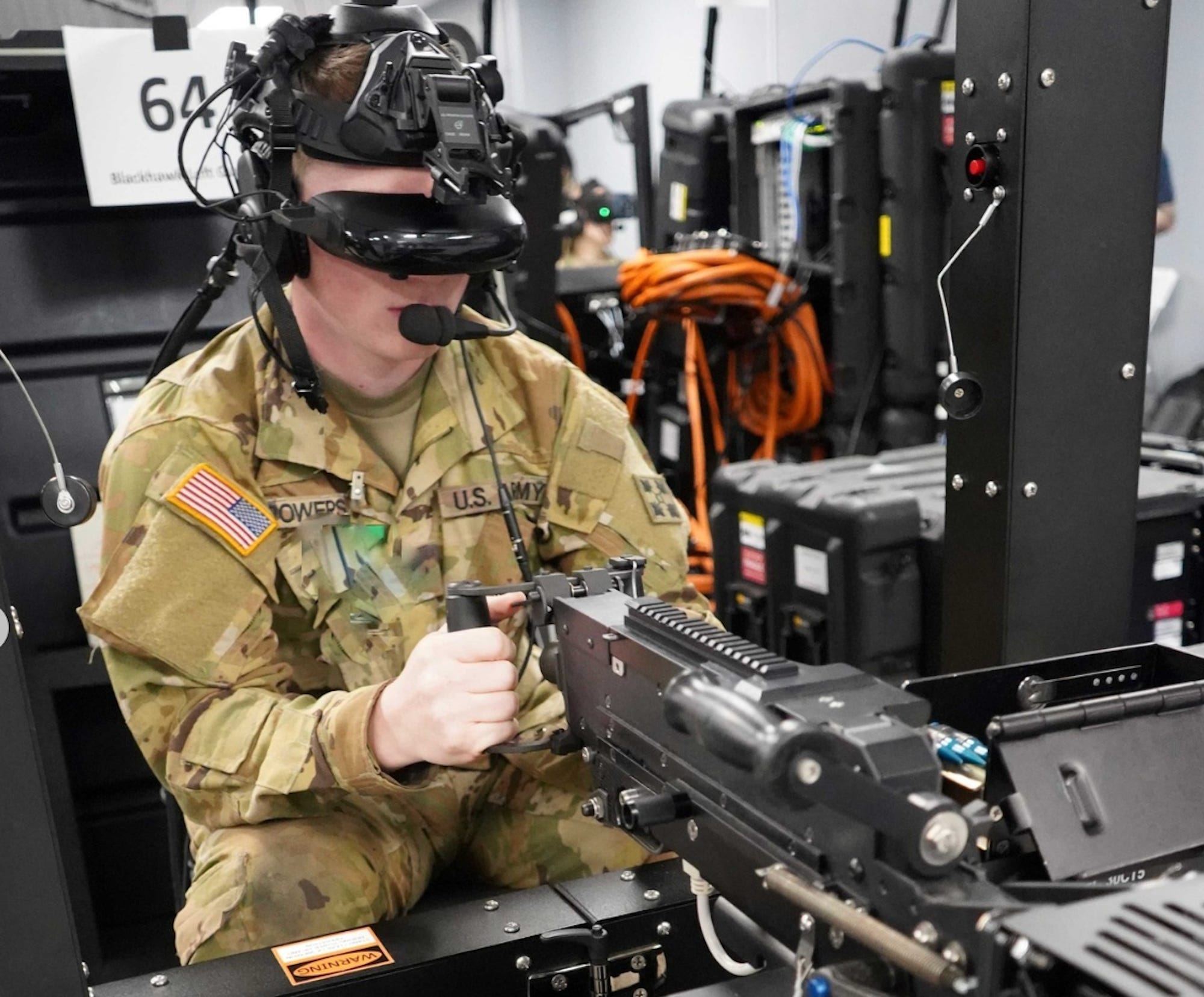 A man wearing camouflage gear with a US flag patch wears a headset that covers his eyes and holds onto a black piece of machinery