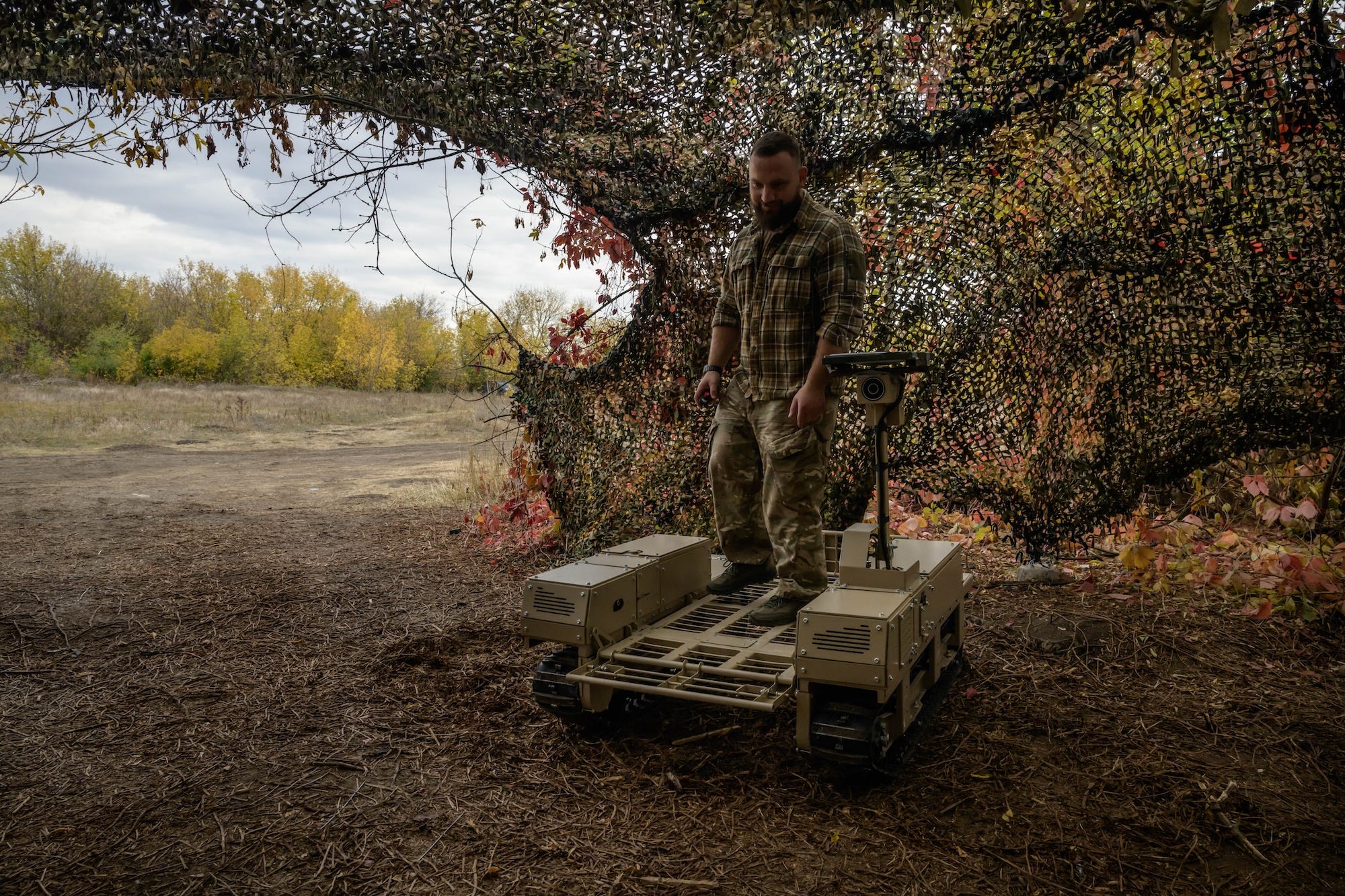 A man in camouflage gear stands outside under a camouflage net with a beige robot on the ground