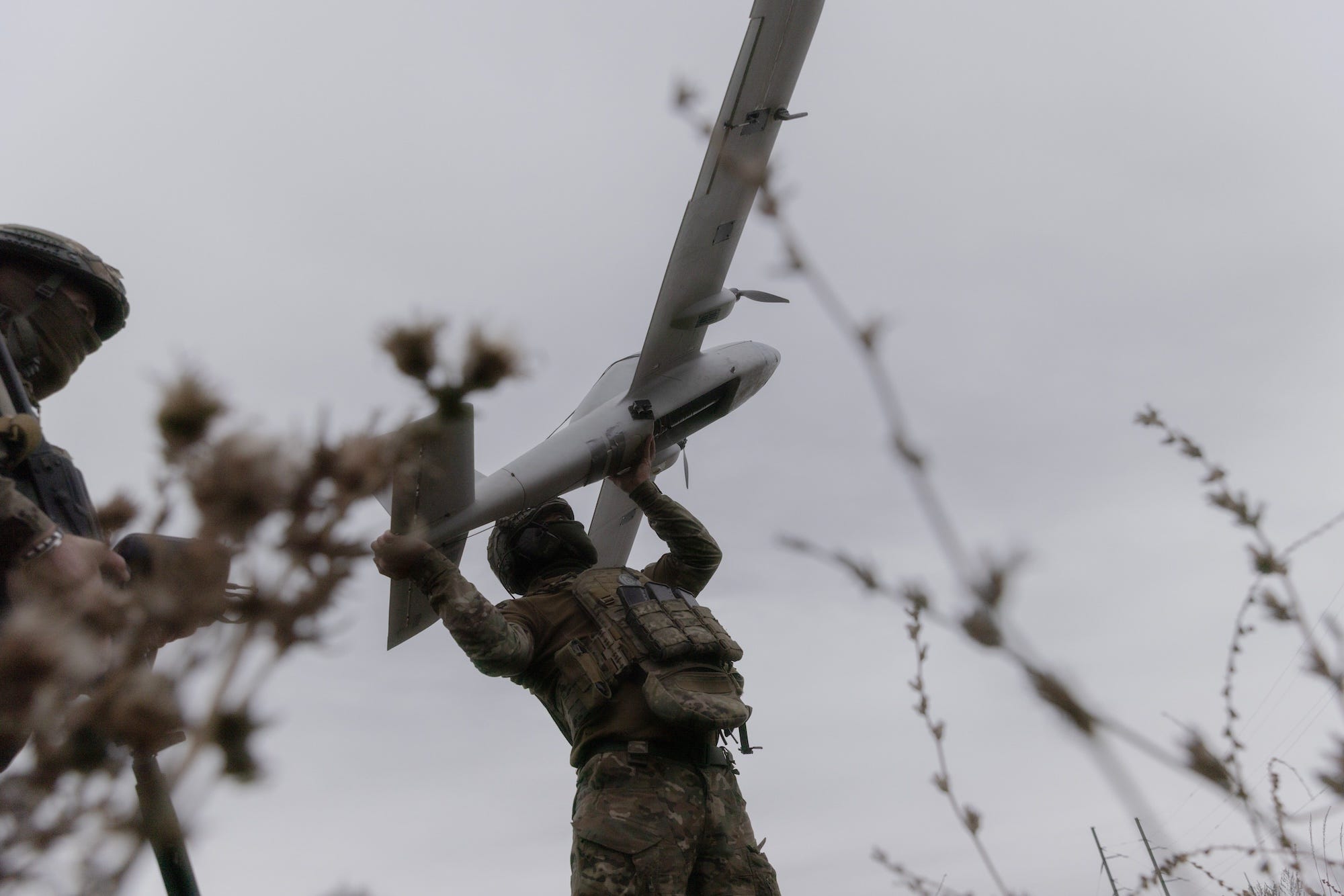 A man in camouflage gear holds a large grey drone in the air under a grey sky