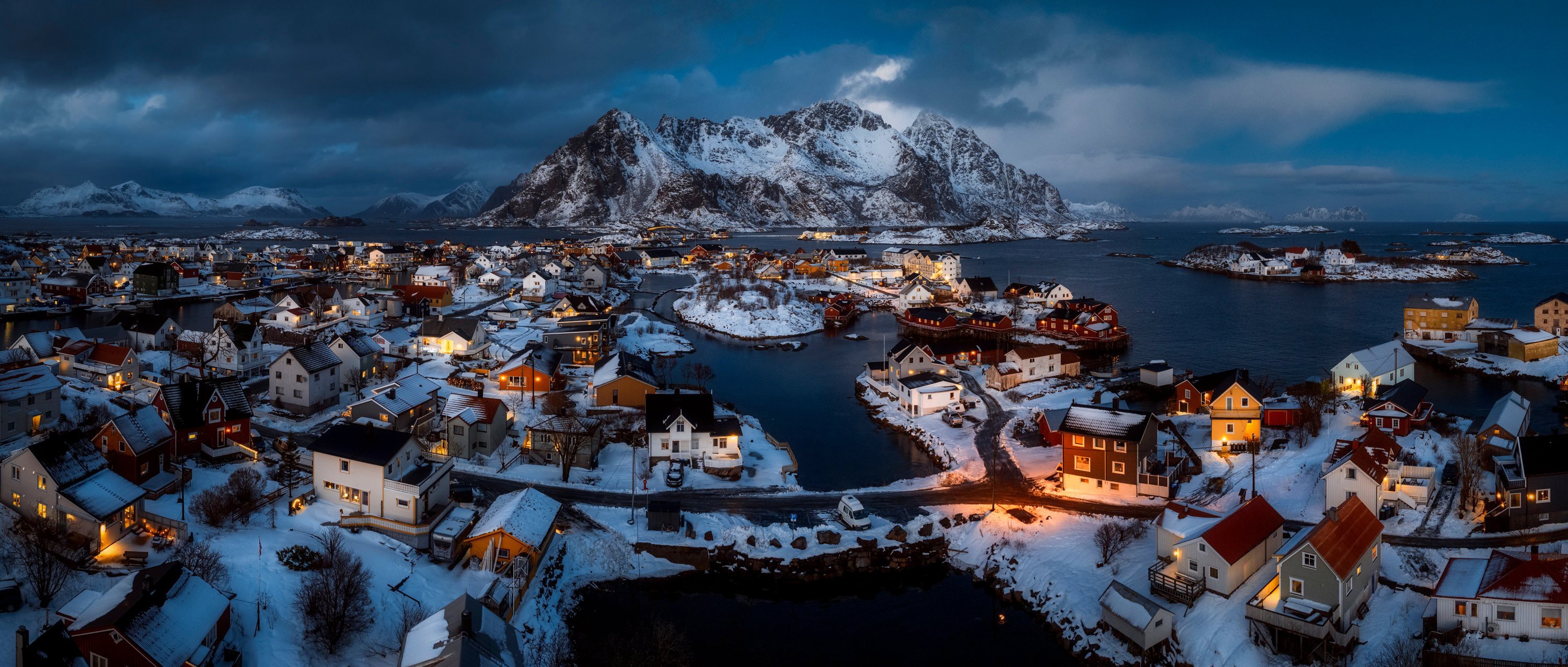 A wide aerial view of the houses of a Norwegian island village, with light snow covering the ground and mountains in the background